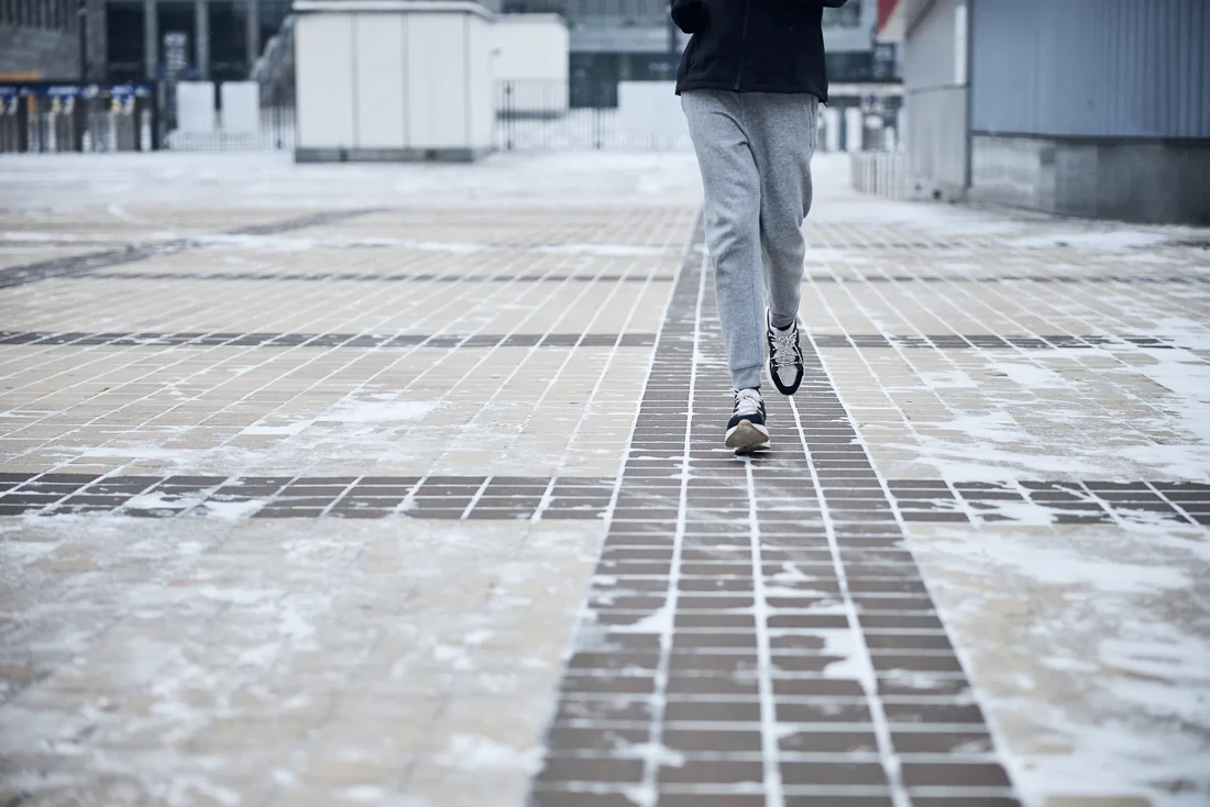 A person walking along a bare, dry paved path running straight through a plaza that is otherwise covered in snow